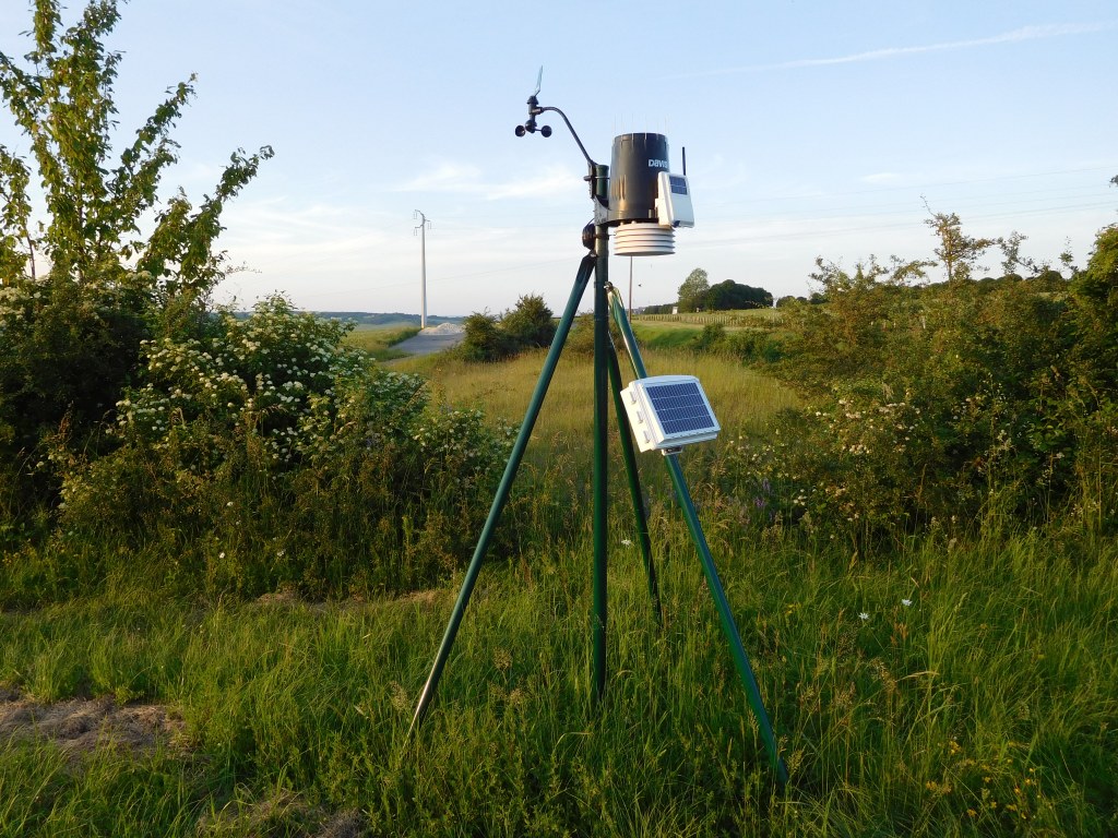 A Davis Vantage Pro wireless weather station on a green tripod in a grassy field: anemometer on a short arm at the top, black rain collector above the white radiation shield, and a small solar-panel box on one leg